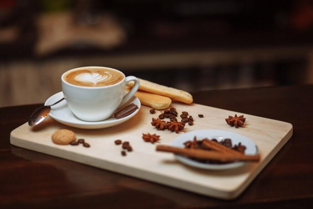 A cappuccino in a white cup sits on a wooden board with cookies, coffee beans, cinnamon sticks, and star anise. - Home Instead