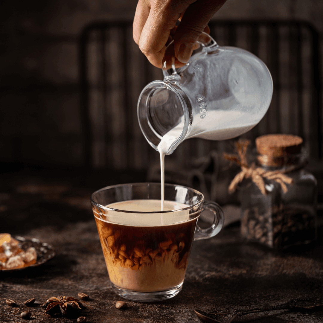 A hand pouring milk from a glass pitcher into a cup of coffee on a rustic table with jars and coffee beans in the background. - Home Instead