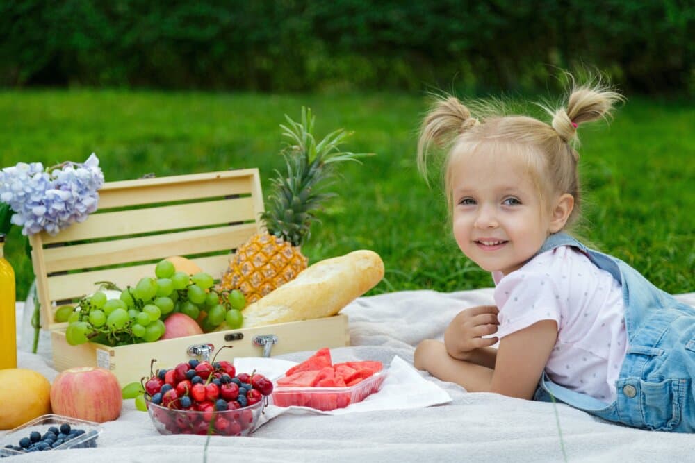 A little girl with pigtails smiles while lying on a picnic blanket with fruits and bread spread out in front of her. - Home Instead