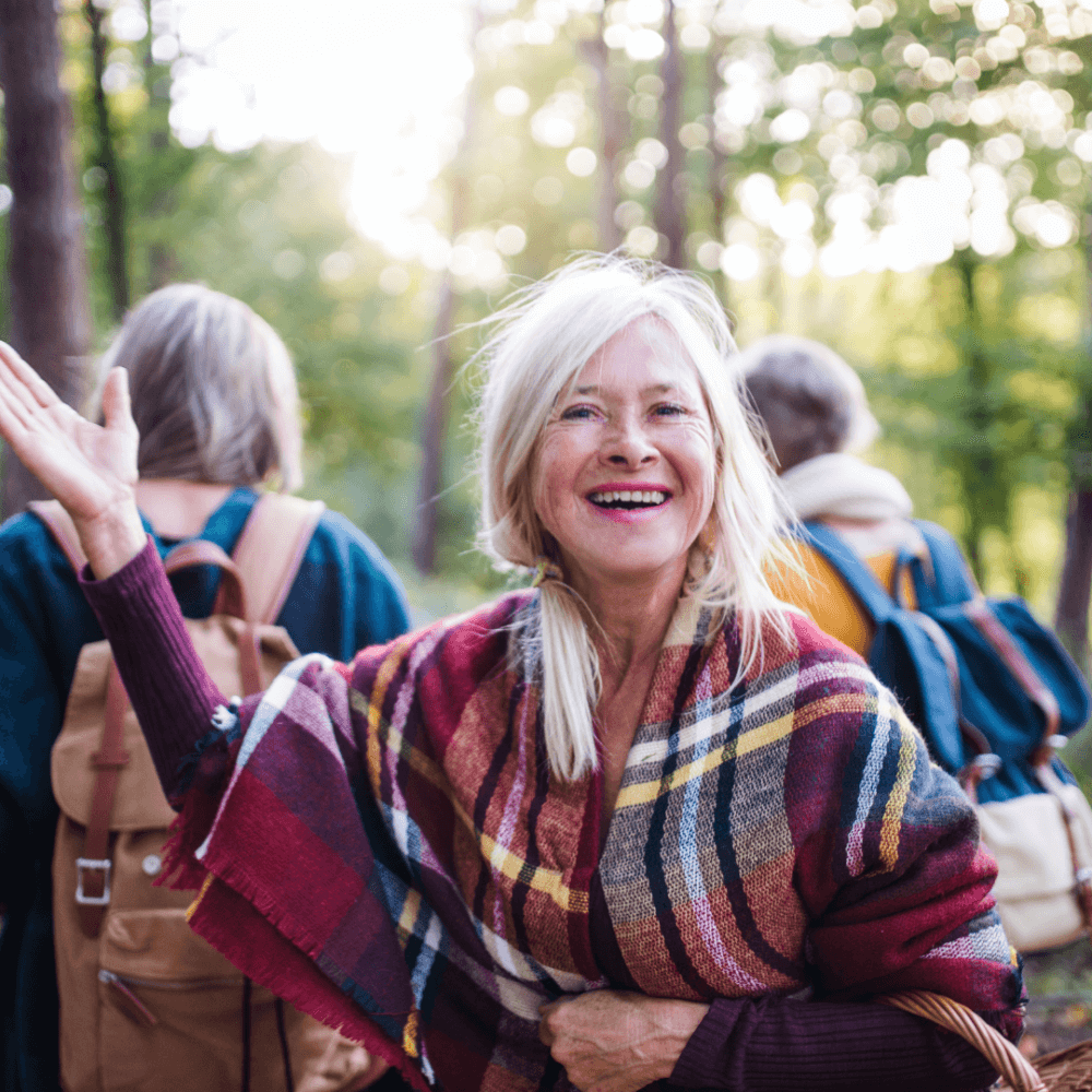 A smiling woman in a plaid shawl waves with her arm up while walking through a forest with others. - Home Instead