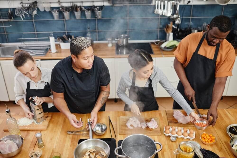 Four people in aprons cooking in a kitchen, chopping ingredients and preparing food on wooden counters. - Home Instead