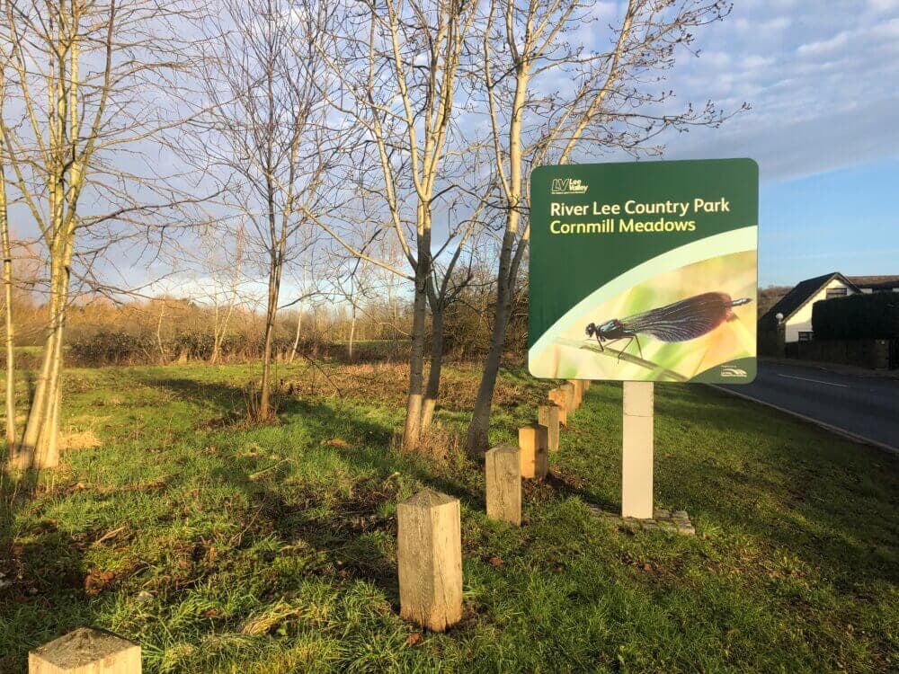 Sign at River Lee Country Park, Cornmill Meadows, with bare trees and grass in the background on a sunny day. - Home Instead