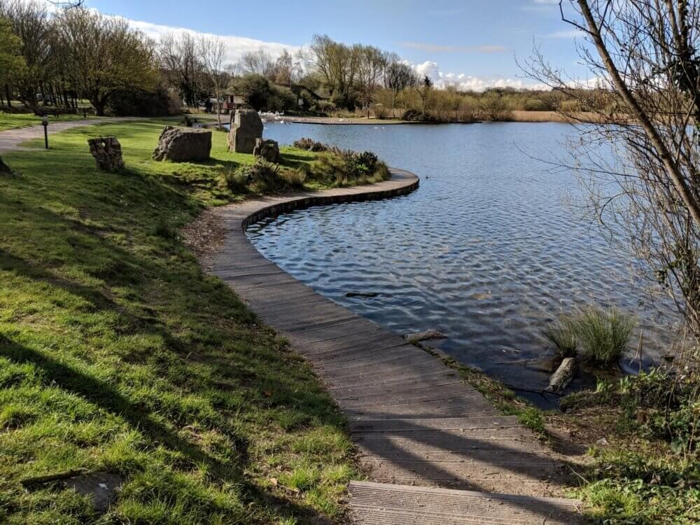 A curved wooden pathway beside a calm lake, surrounded by grass and trees under a blue sky with scattered clouds. - Home Instead