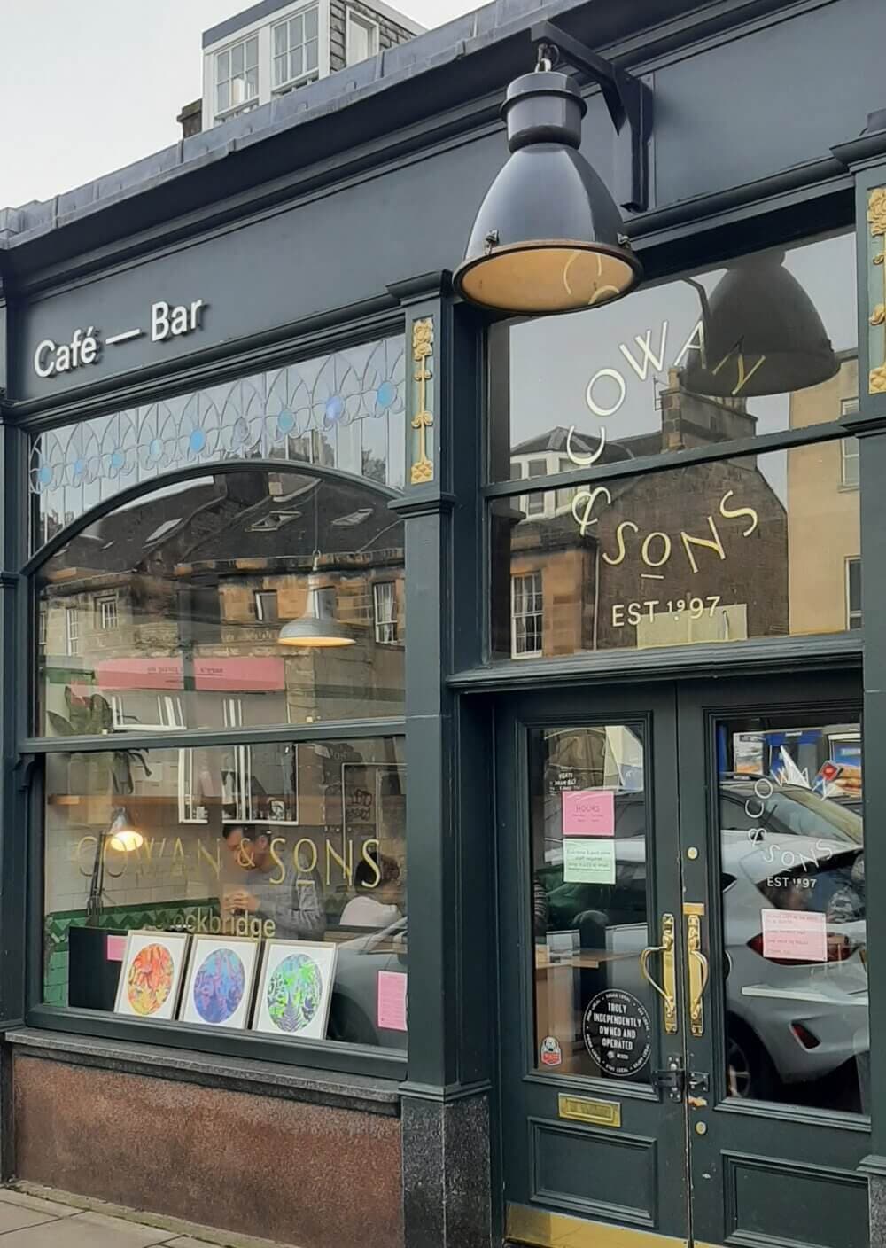 Storefront of Cowan & Sons Café — Bar with large windows displaying artwork and signage indicating establishment in 1997. - Home Instead