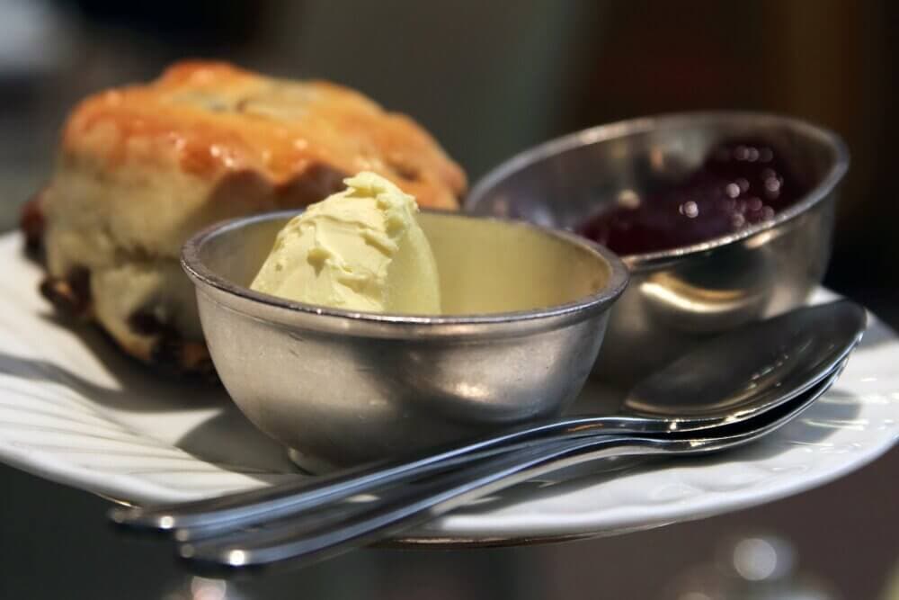 A plate with a scone, a dish of clotted cream, a dish of jam, and two spoons. - Home Instead