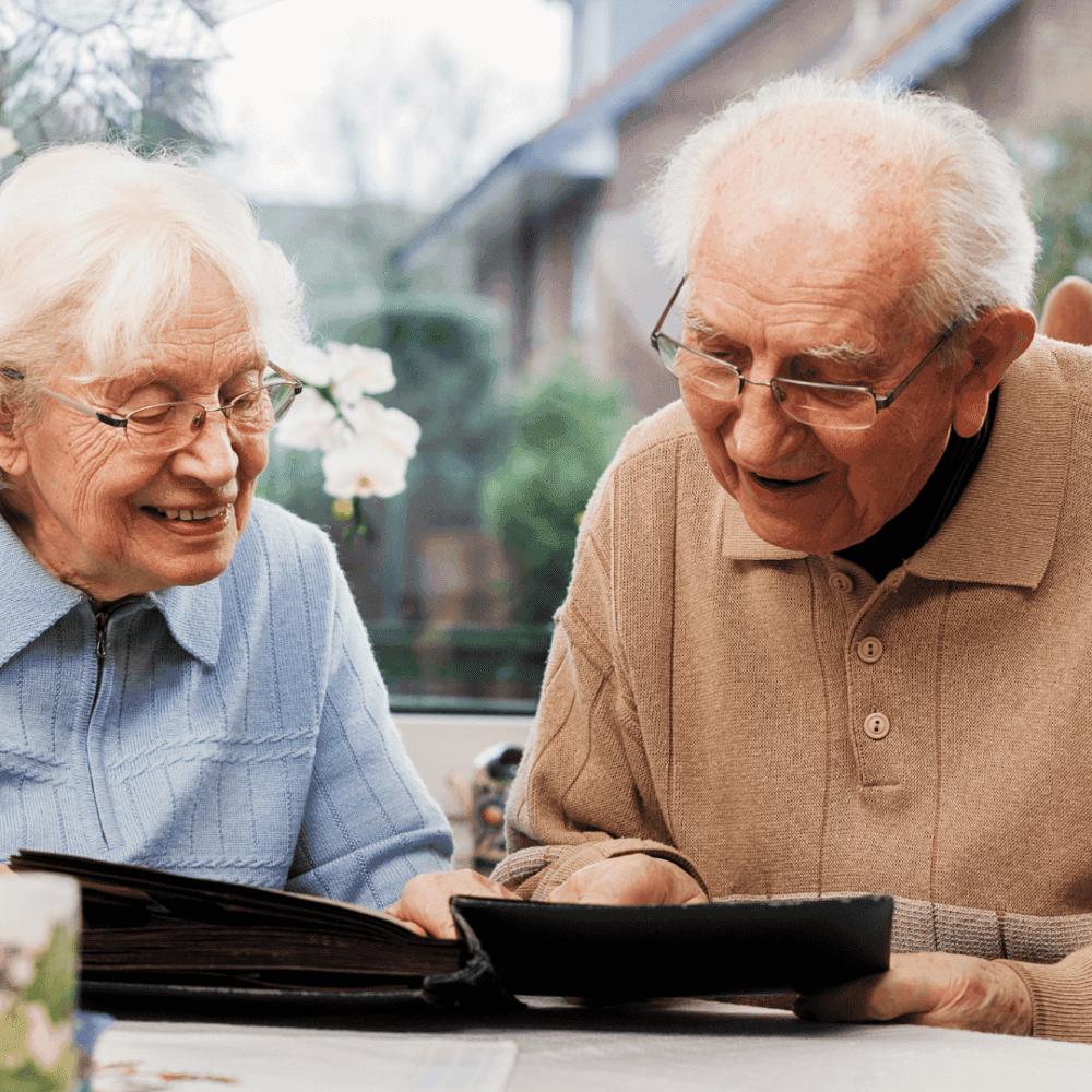 Two elderly people smiling and looking at a photo album together at a table, with a homey background. - Home Instead