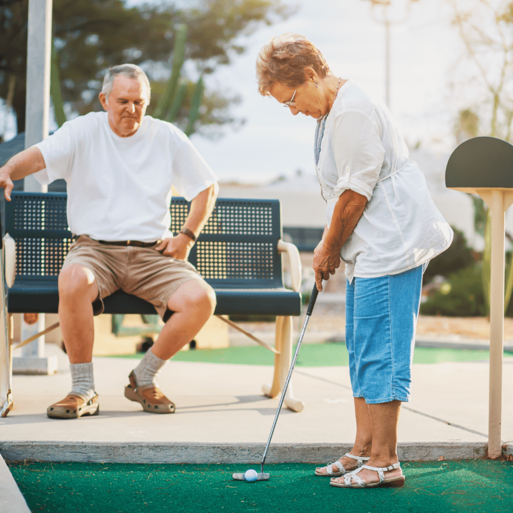 An older woman putts a golf ball while an older man sits on a bench watching her. - Home Instead