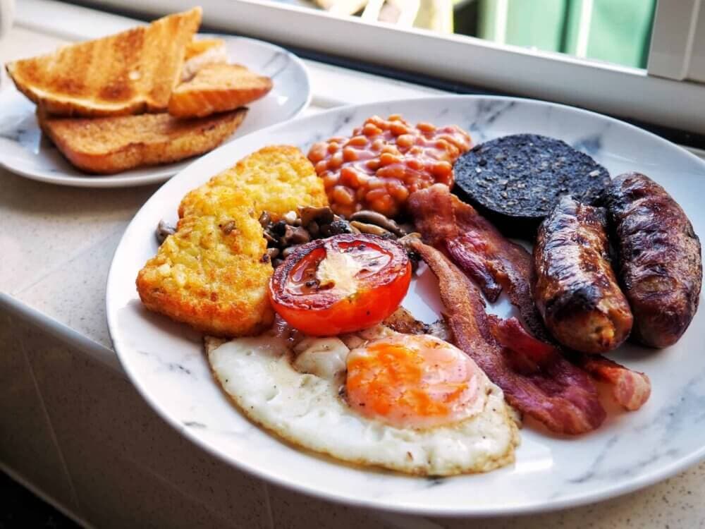A hearty breakfast plate with fried eggs, sausages, bacon, black pudding, beans, tomatoes, mushrooms, and hash browns. - Home Instead