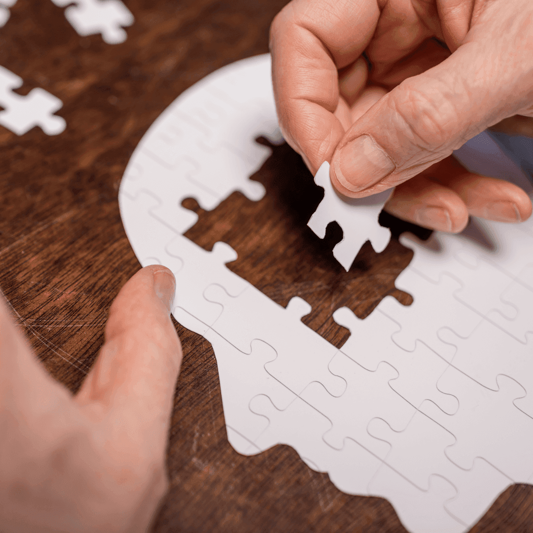 Hands assembling a jigsaw puzzle in the shape of a human head on a wooden surface. - Home Instead