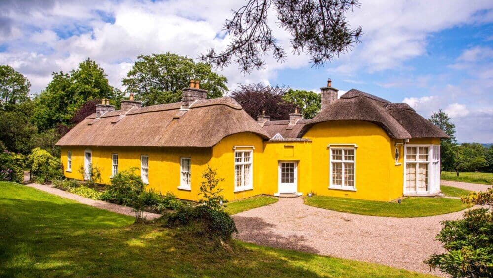 Yellow thatched-roof cottage with white-framed windows, surrounded by green trees and a gravel path, on a sunny day. - Home Instead