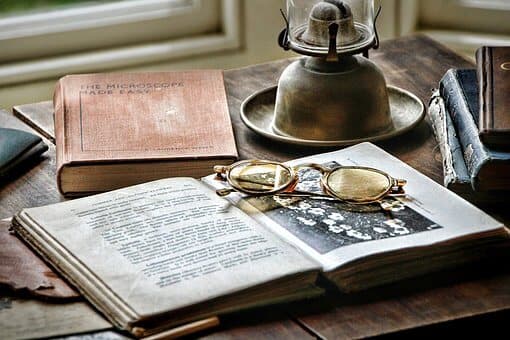 Open book with glasses on top, surrounded by other books and a vintage oil lamp on a wooden table near a window. - Home Instead