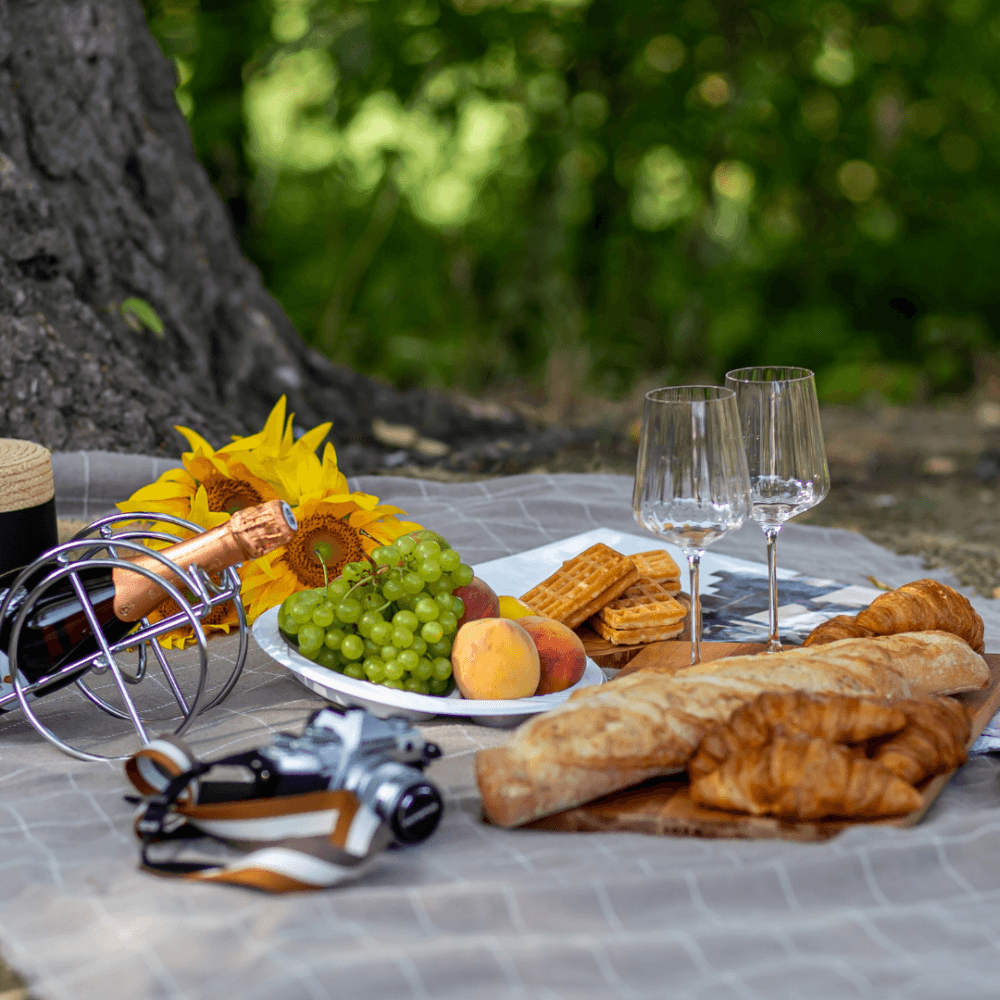 Picnic setup with croissants, grapes, peaches, waffles, wine, glasses, a camera, sunflowers, and a nature backdrop. - Home Instead