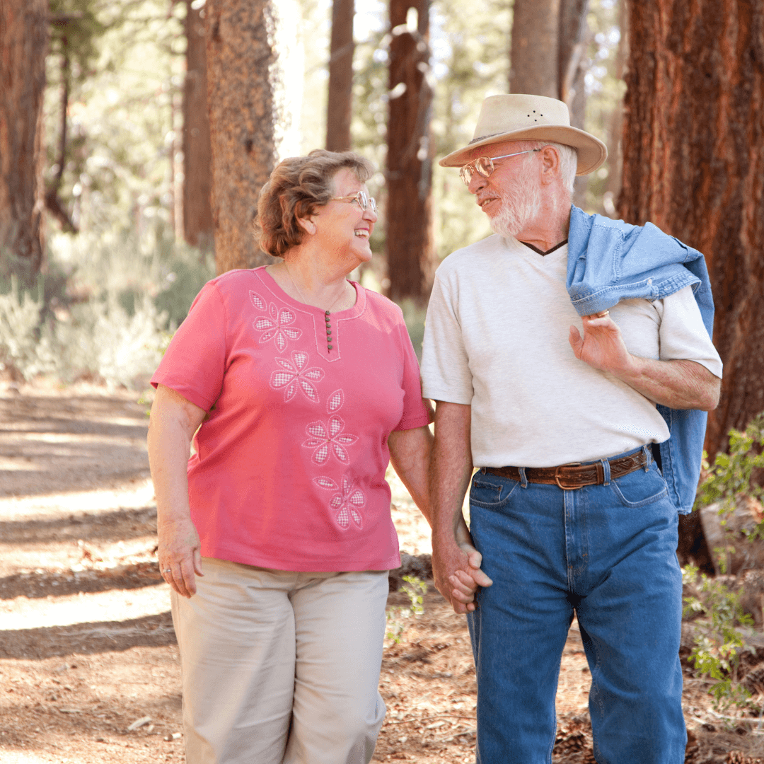 An elderly couple walks hand in hand through a forest, smiling at each other affectionately. - Home Instead