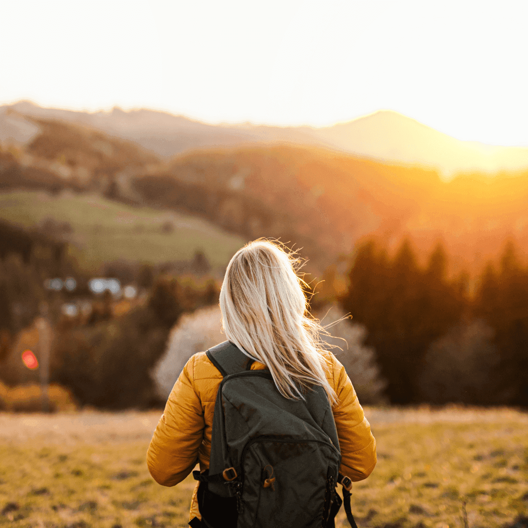 A person with a backpack watches the sunrise over hilly countryside with trees and fields. - Home Instead
