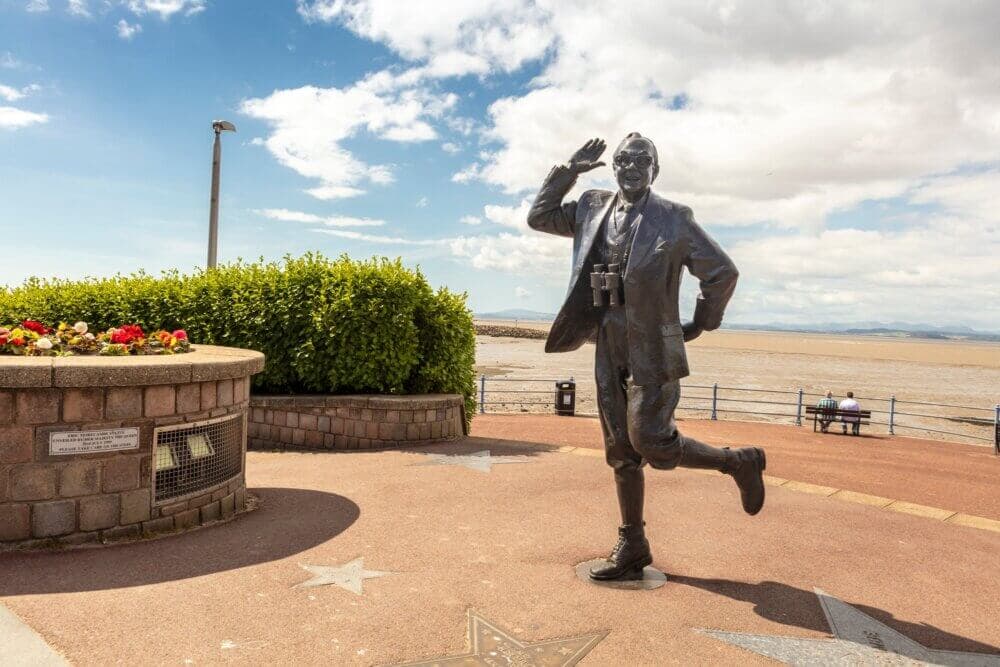 Bronze statue of a man in a suit with binoculars around his neck, posed mid-stride near the coast on a sunny day. - Home Instead