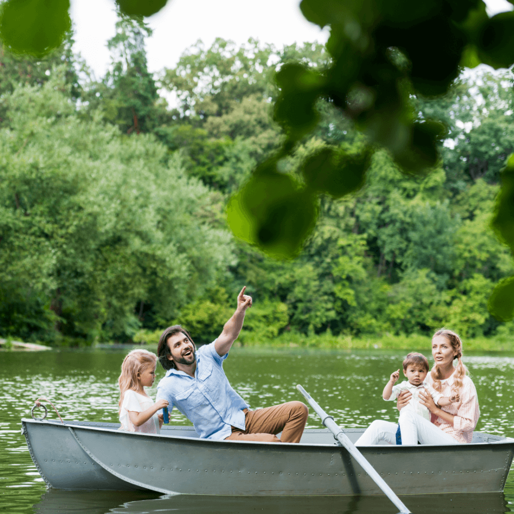 A family of four enjoys a boat ride on a serene lake, surrounded by lush greenery. The father points at something above. - Home Instead