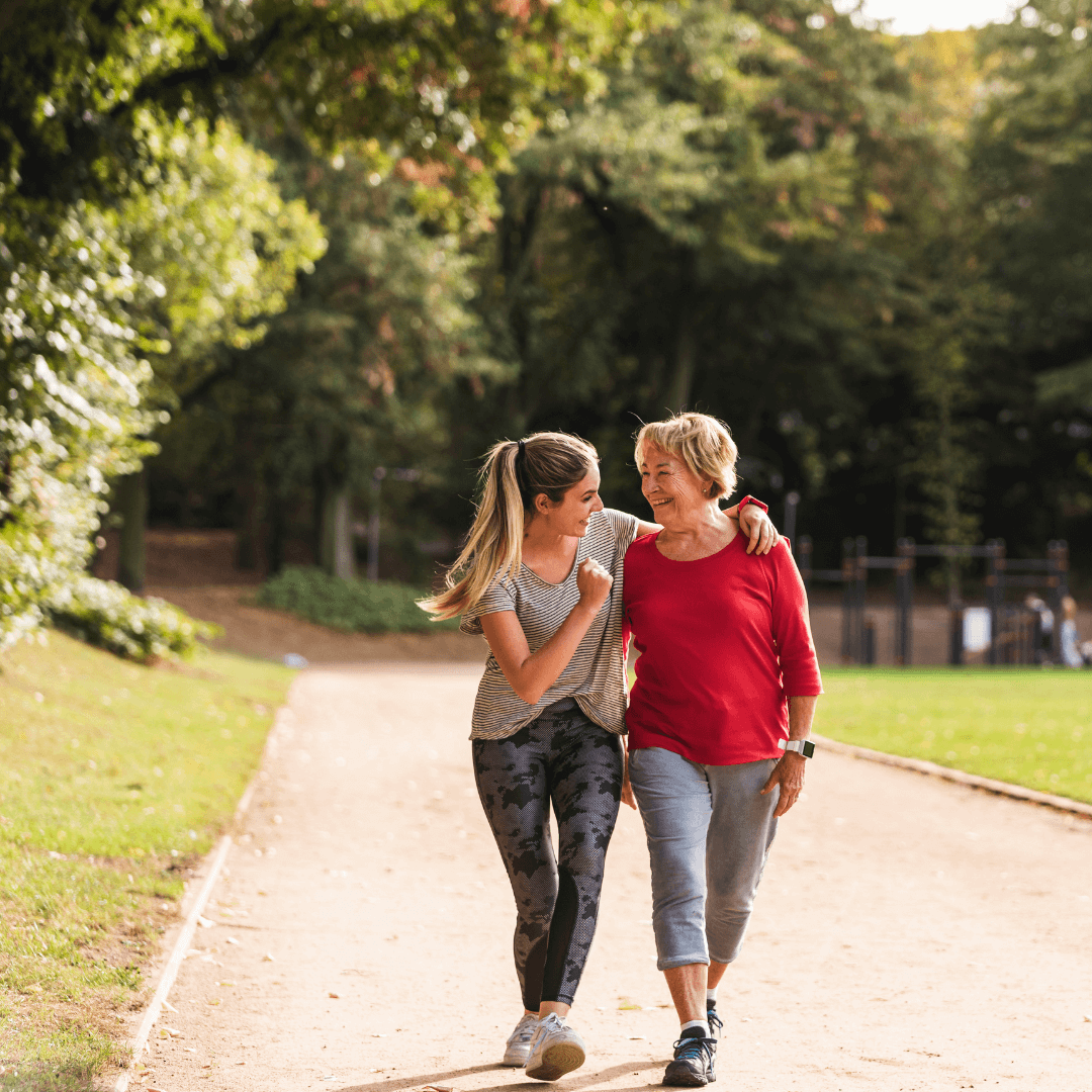 A younger woman and an older woman walk together on a park path, smiling and talking. - Home Instead