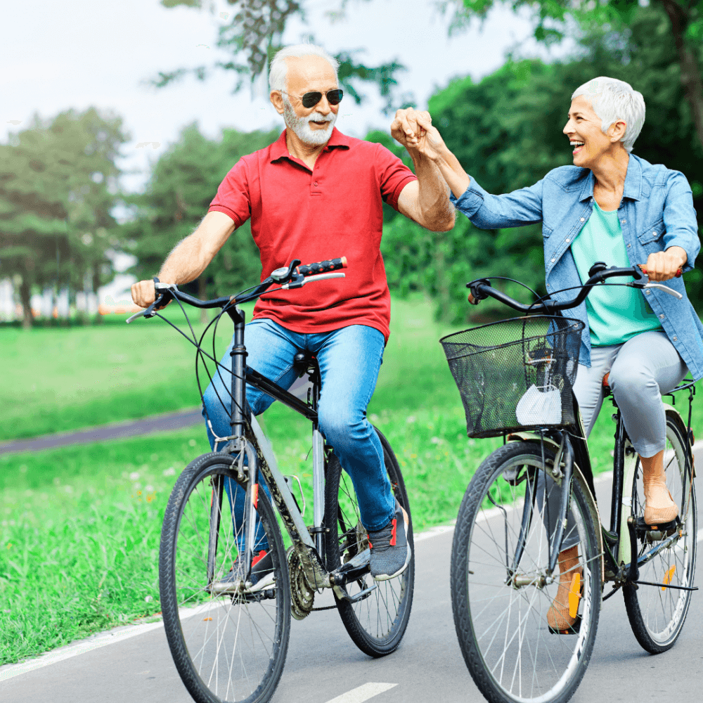 Elderly couple riding bicycles on a path, holding hands and smiling, with trees and greenery in the background. - Home Instead