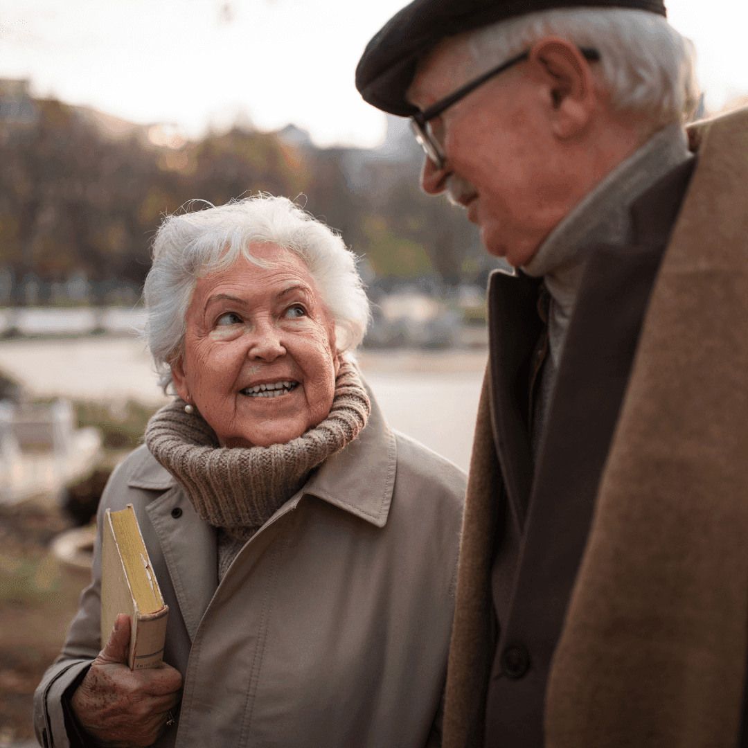 A smiling elderly couple, wearing coats, holding a book while walking outdoors on a cloudy day. - Home Instead
