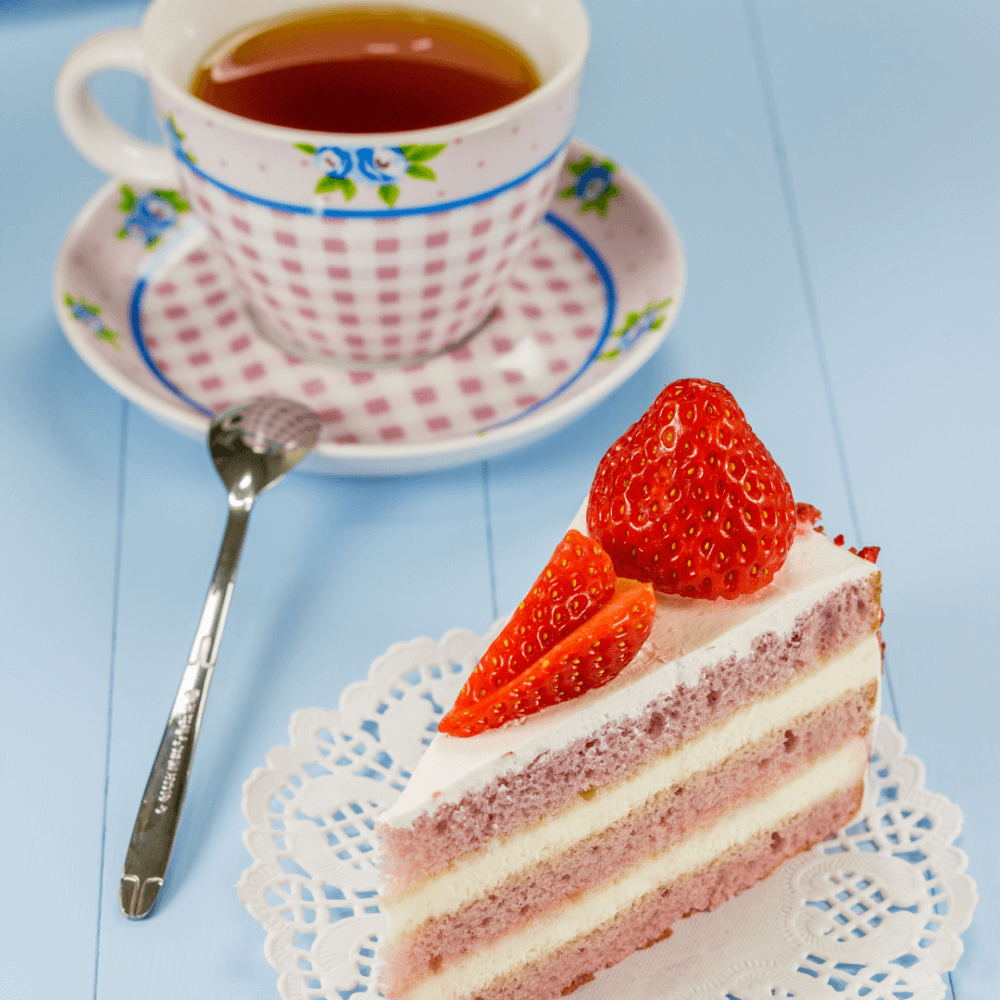 A slice of strawberry cake topped with fresh strawberries on a lace doily, next to a teacup on a saucer with tea. - Home Instead