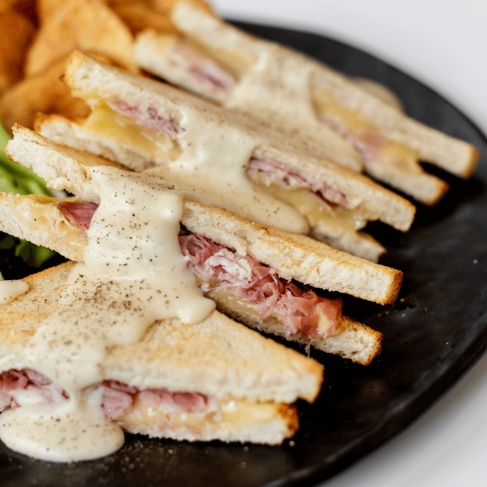 Close-up of toasted ham and cheese sandwiches topped with melted cheese and black pepper on a dark plate. - Home Instead