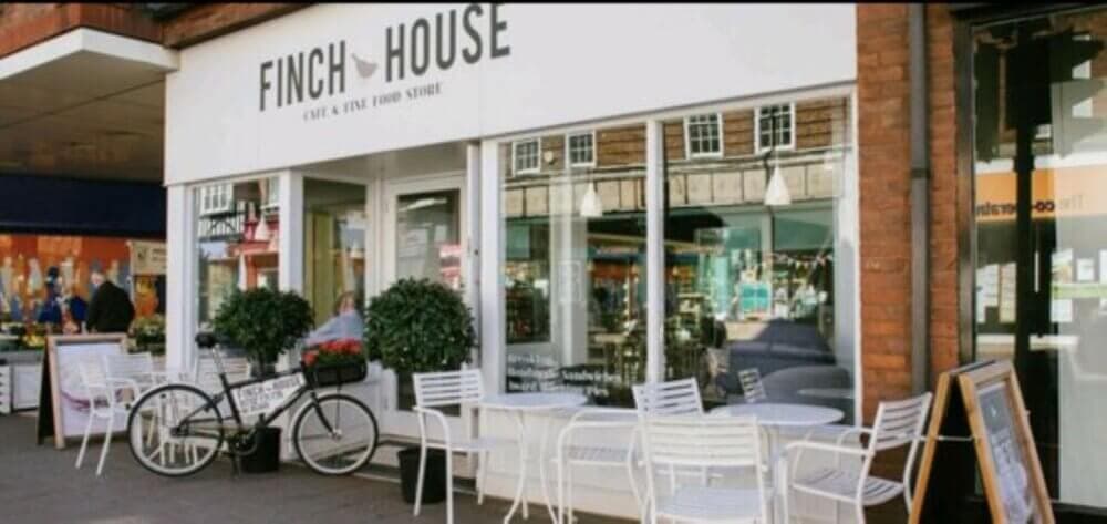 Street view of Finch House cafe with outdoor seating, white chairs and tables, a black bicycle, and potted plants. - Home Instead
