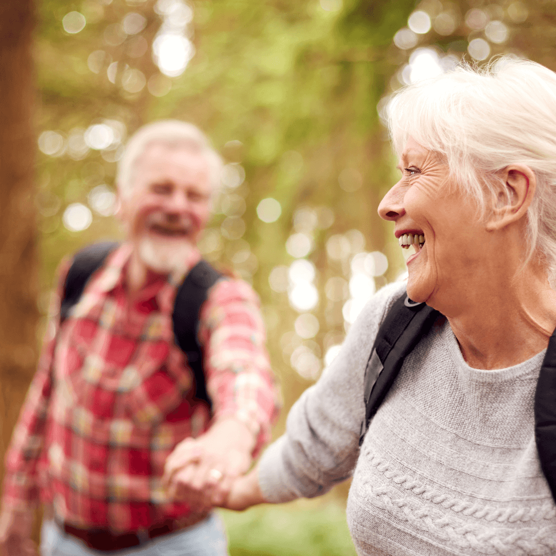 Elderly couple with backpacks smile and hold hands while walking through a sunlit forest. - Home Instead