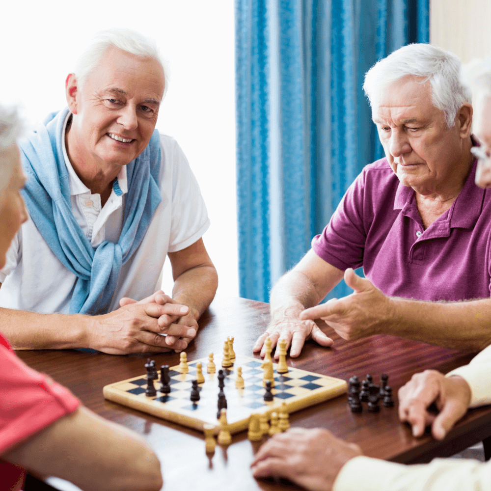 Four elderly men playing chess at a table, with one man observing and others focused on the game. - Home Instead