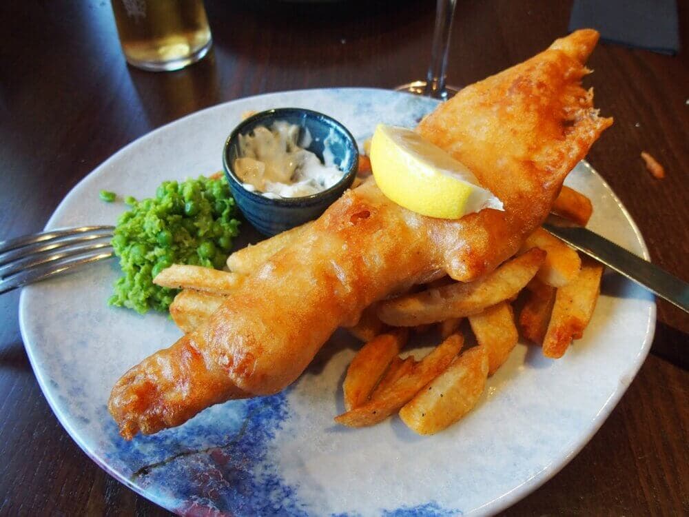 A plate of fish and chips with a lemon wedge, mushy peas, and a small cup of tartar sauce on a dark wooden table. - Home Instead