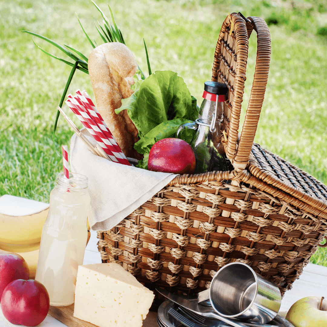 Picnic basket with bread, lettuce, red apples, bottle of lemonade, cheese, and utensils on a table outdoors. - Home Instead