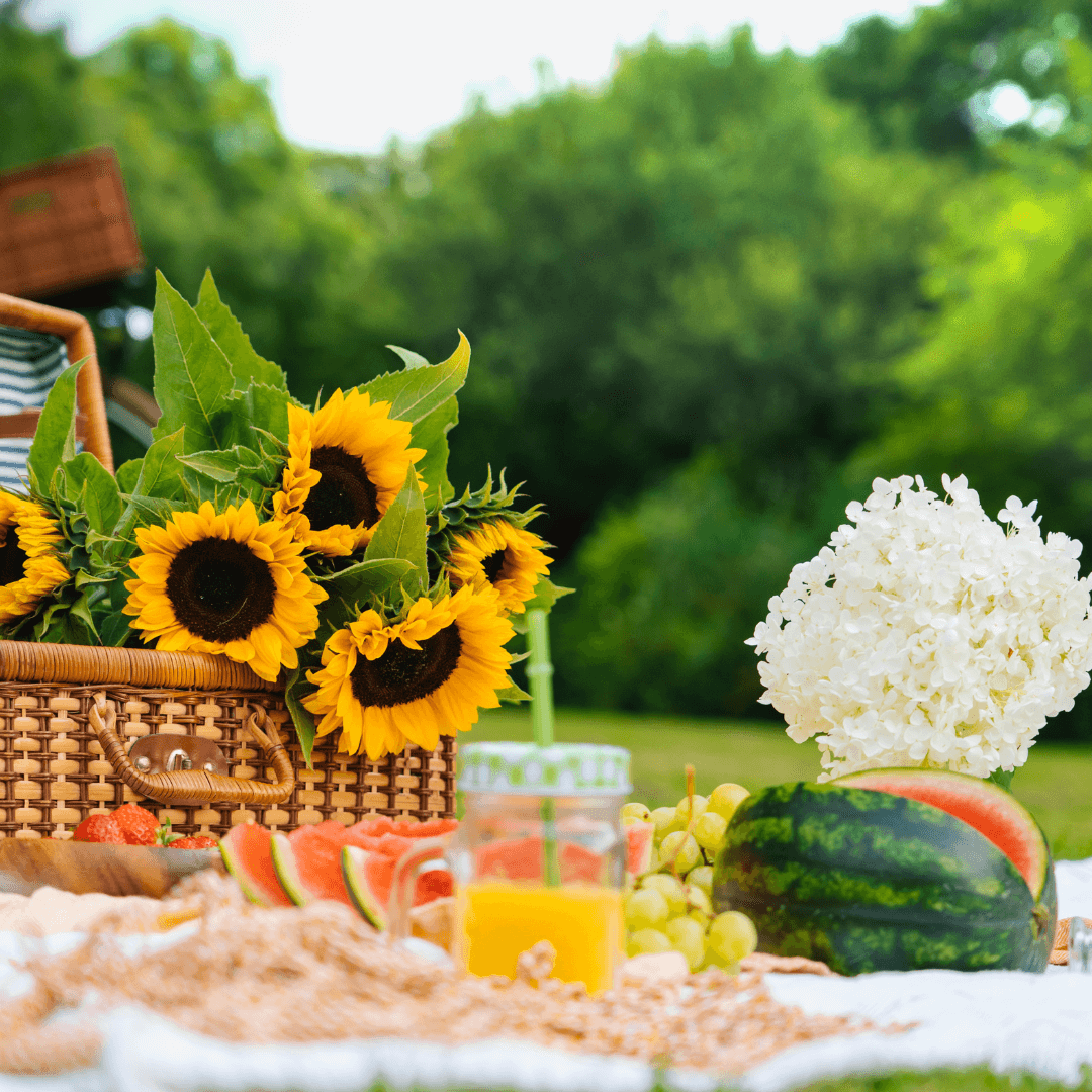A picnic setup with a basket of sunflowers, a white hydrangea, various fruits, and a drink on a blanket outdoors. - Home Instead