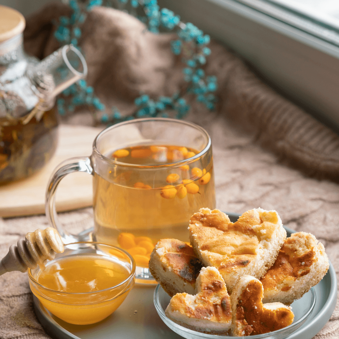 A cozy setting with heart-shaped pastries on a plate, a glass of tea, a honey jar with a dipper, and a teapot in the background. - Home Instead