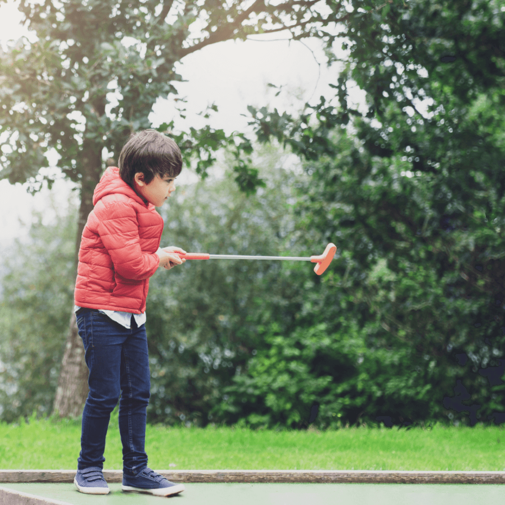 A young boy in a red jacket plays miniature golf outdoors, preparing to hit the ball with a putter. - Home Instead