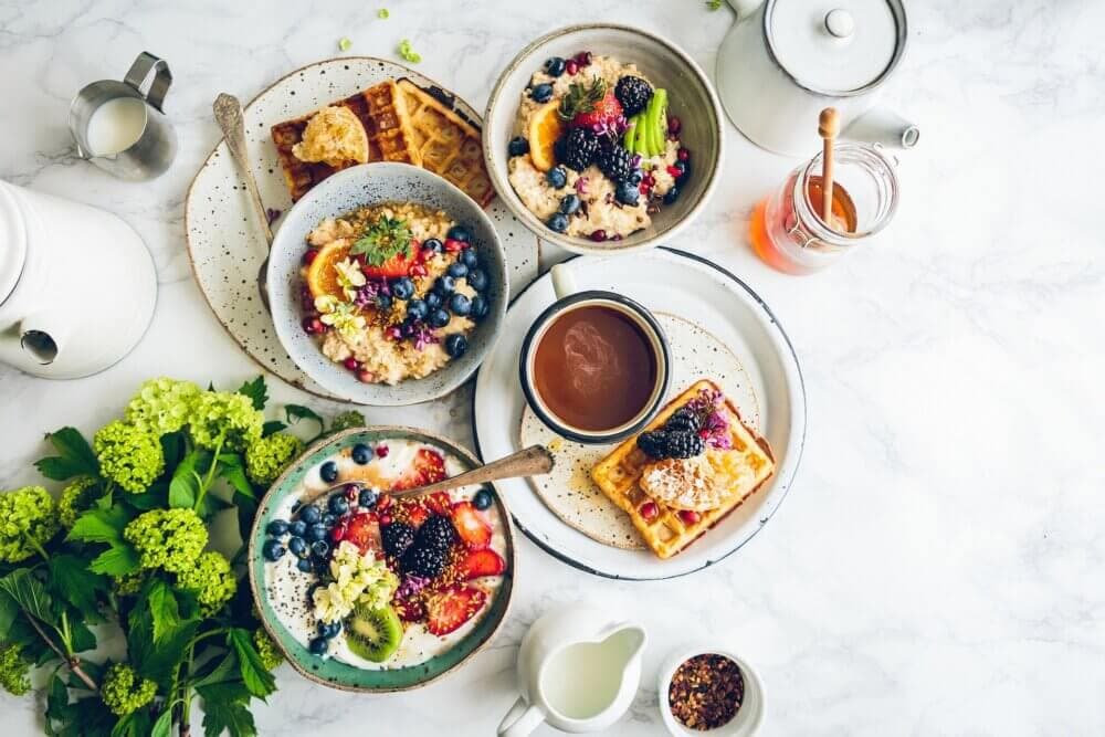 A breakfast spread with waffles, oatmeal bowls, fruits, a cup of coffee, and a pot of honey on a marble table. - Home Instead