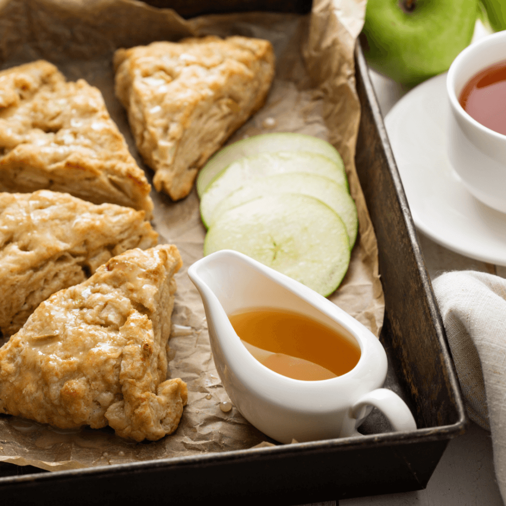 Tray of scones with sliced green apples, a white pitcher of honey, and a cup of tea on a white saucer. - Home Instead
