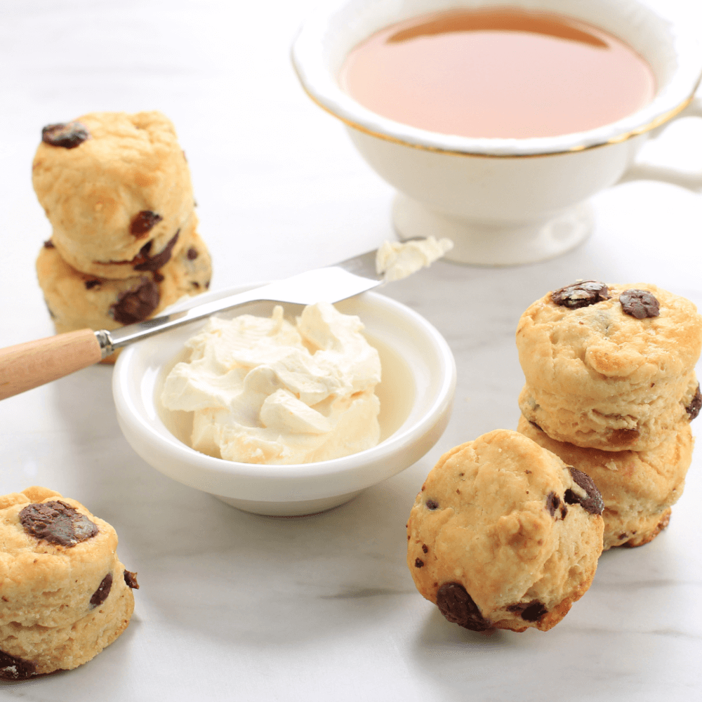 Scones with chocolate chips next to a bowl of cream and a cup of tea on a white surface. - Home Instead