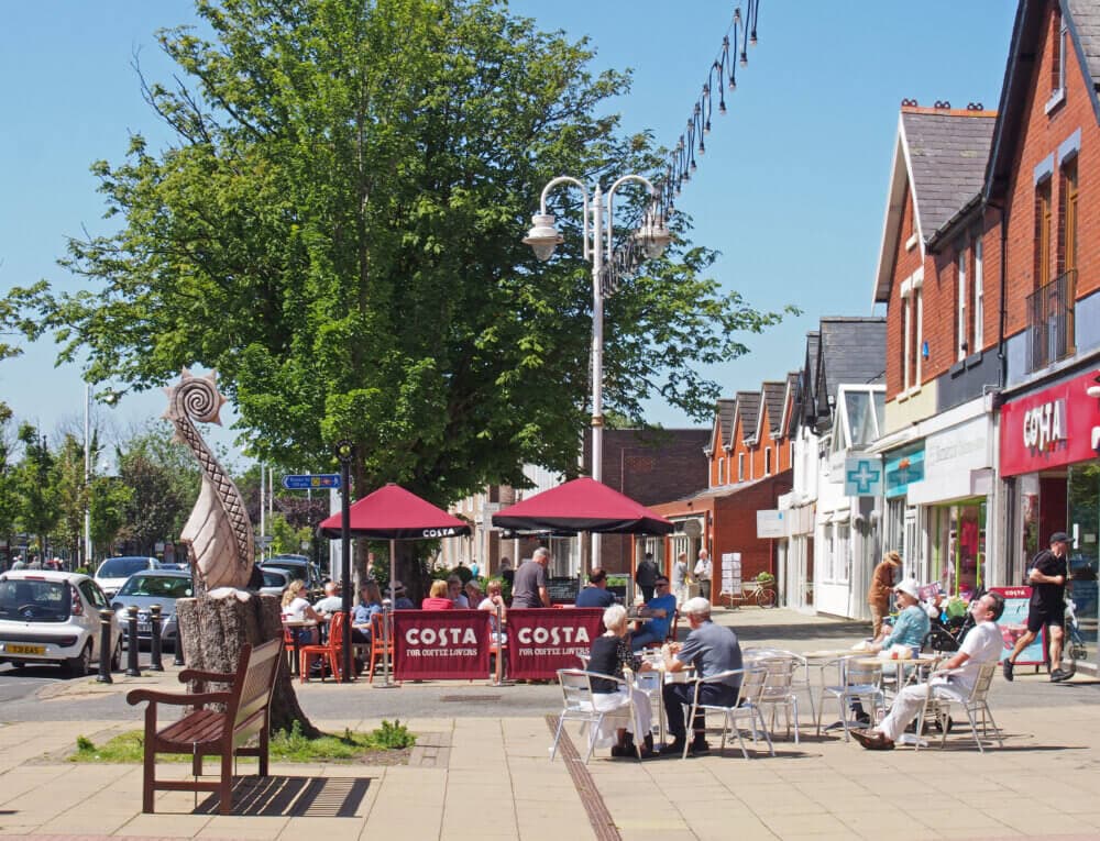 Street with outdoor café seating, people dining, red brick buildings, shops, a tree, and a public art sculpture. - Home Instead