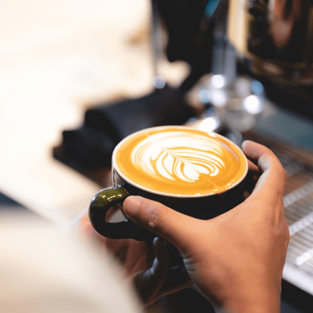 A person holds a cup of coffee with intricate latte art, near an espresso machine. - Home Instead