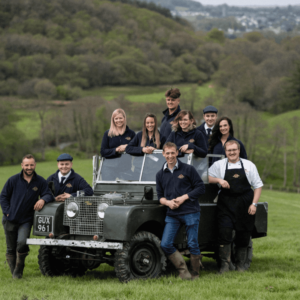 Group of people posing around a vintage Land Rover in a grassy field with hills and trees in the background. - Home Instead