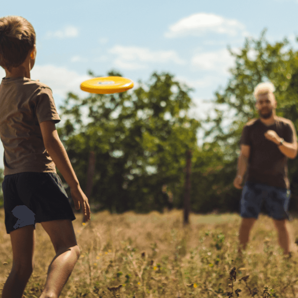 A child prepares to catch a yellow frisbee thrown by an adult in a sunny outdoor setting with trees and grassy field. - Home Instead