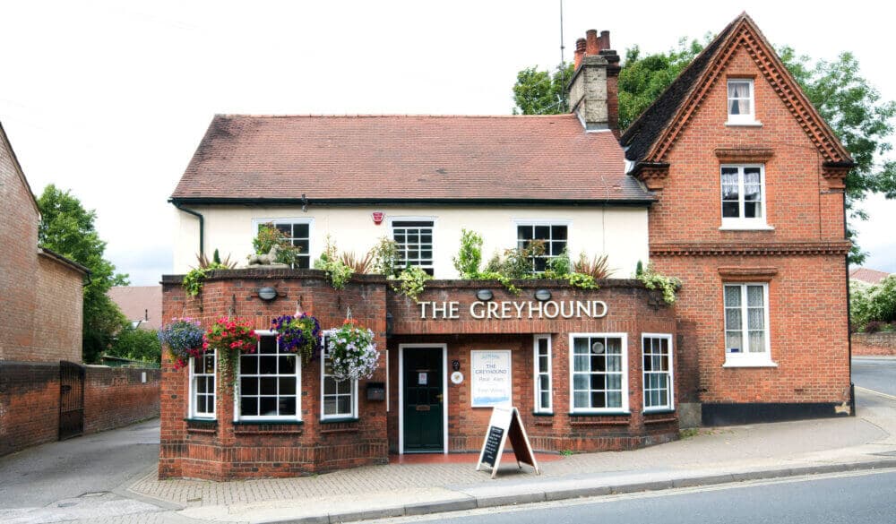 A traditional brick building with a sign that reads "The Greyhound"; a pub entrance with hanging flowers and a sidewalk board. - Home Instead