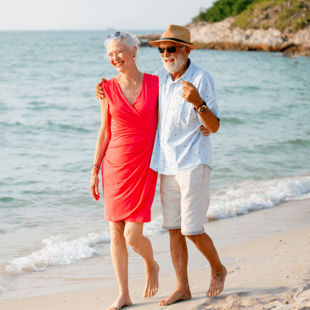 An elderly couple, dressed in summer clothes, happily walking barefoot on a beach with the sea in the background. - Home Instead
