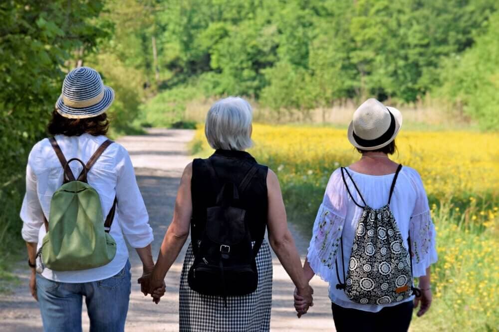 Three women wearing hats and backpacks walk hand in hand down a path through a green forest with yellow flowers. - Home Instead
