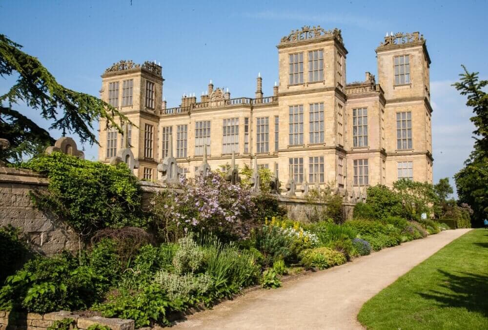 A large historic stone mansion with a garden path and lush greenery on a clear, sunny day. - Home Instead