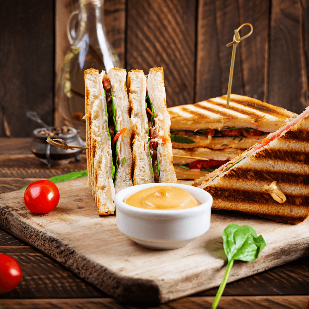 Grilled sandwich quarters stuffed with lettuce, tomatoes, and bacon on a wooden board with a small bowl of dipping sauce. - Home Instead