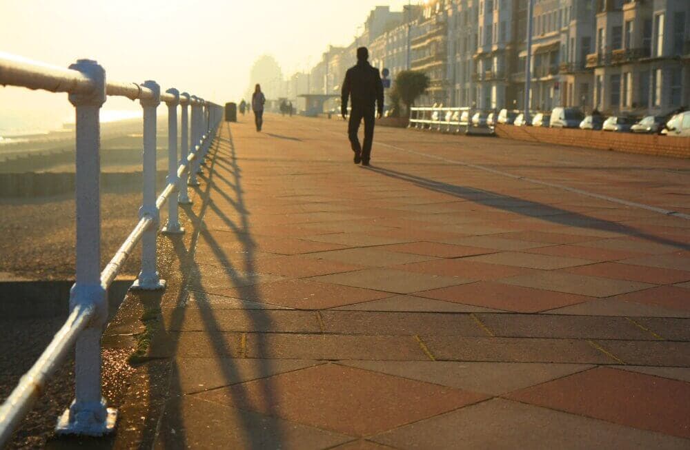 A person walks along a seaside promenade at sunset, with railings and buildings casting long shadows. - Home Instead