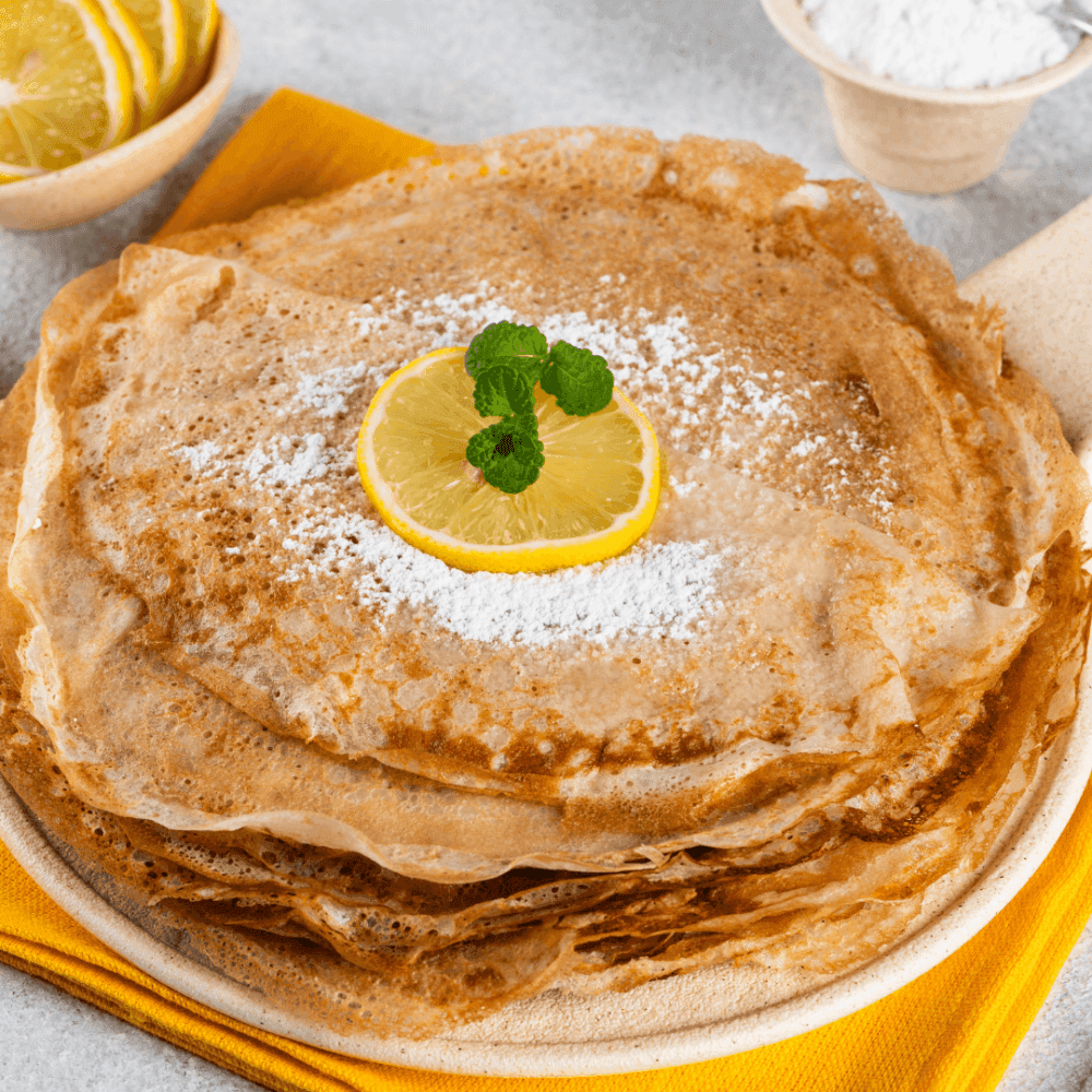 A stack of crepes garnished with a lemon slice and mint leaves, dusted with powdered sugar, on a plate with a yellow napkin. - Home Instead