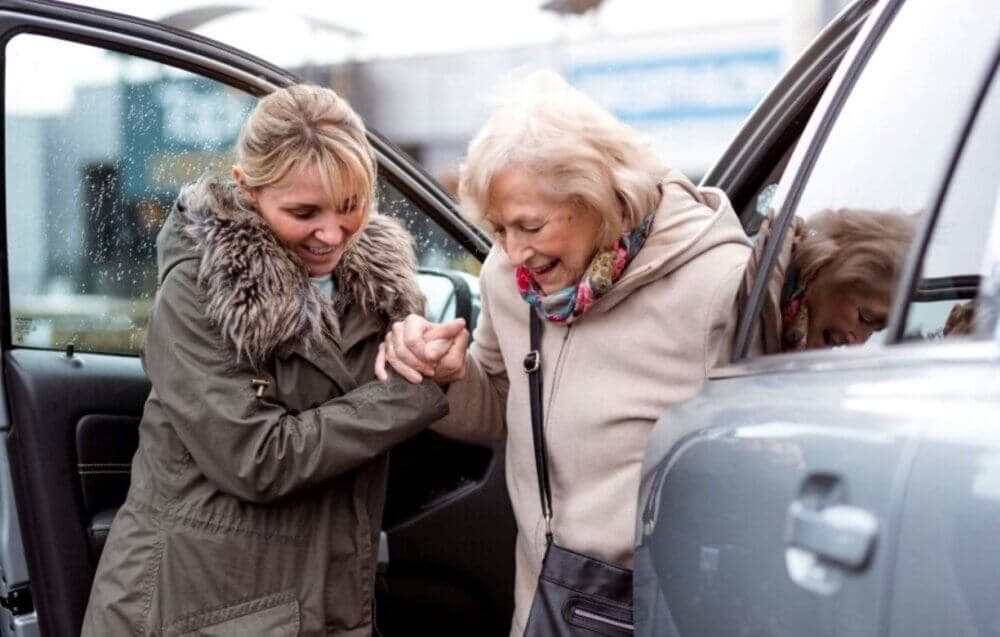 A younger woman helps an elderly woman out of a car on a rainy day, both smiling warmly. - Home Instead