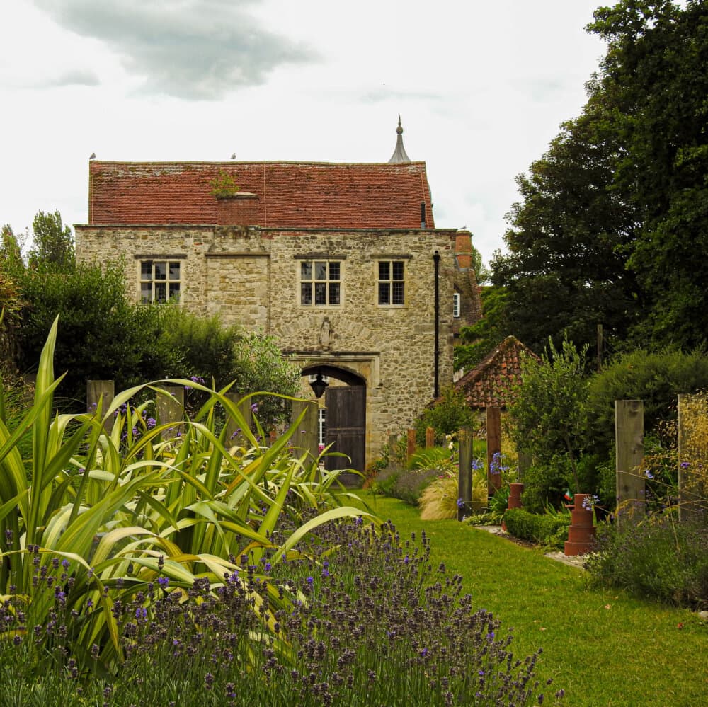 Stone building with a red roof set against a cloudy sky, surrounded by lush greenery and blooming lavender. - Home Instead