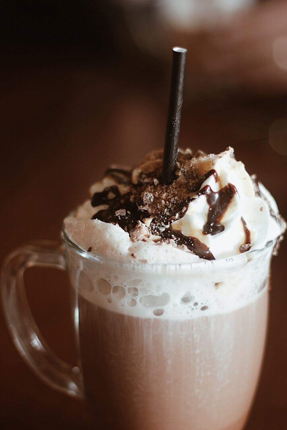 A glass mug filled with hot chocolate, topped with whipped cream, chocolate syrup, and a black straw. - Home Instead